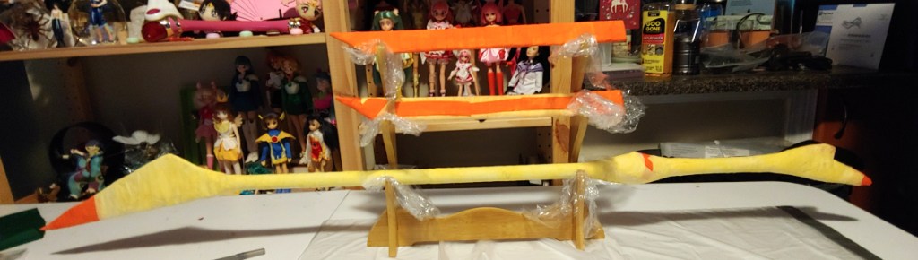 Two swords and a stick drying on a sword holder I use to display finished props. The two swords are covered mostly in orange fabric while the stick is covered mostly in light yellow.
