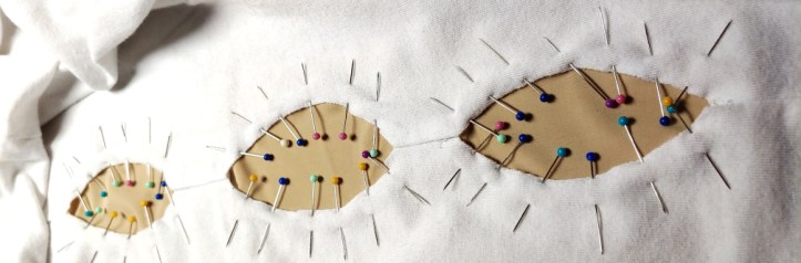 Three cutouts with beige spandex sitting behind them, all pinned along the white edge of the fabric with tiny pins