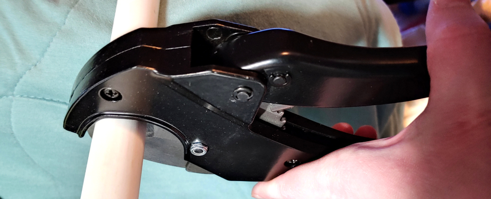 Cutting the pipe with ratcheting cutters. Black metal cutters surround a white PVC pipe to slice it apart!