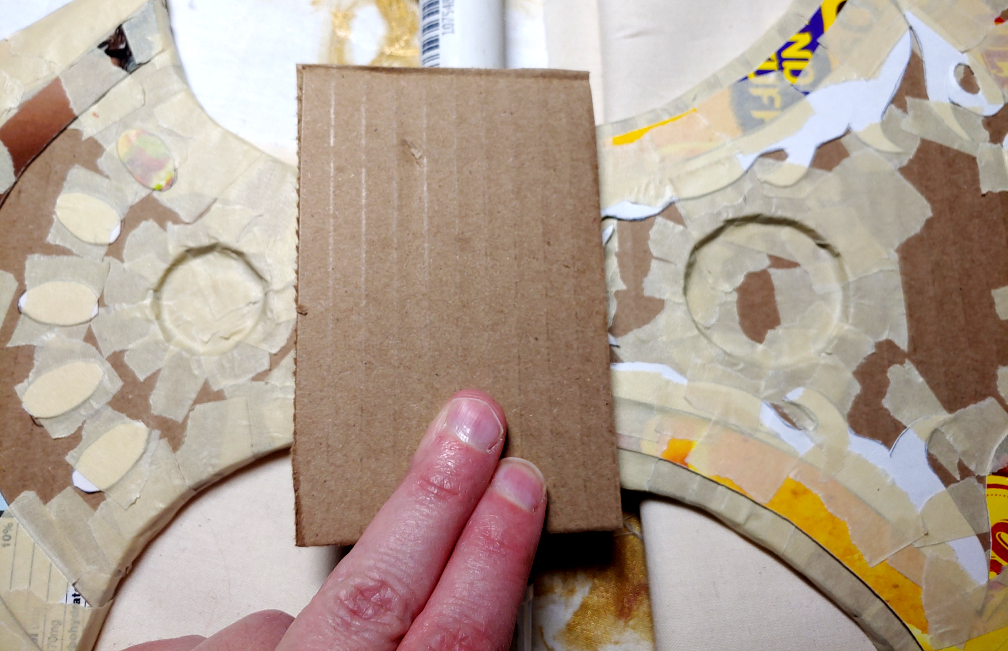 Cutting the main shape of the stabilization box. A large cardboard rectangle placed over the axe heads and pipe, overlapping both to join them together.