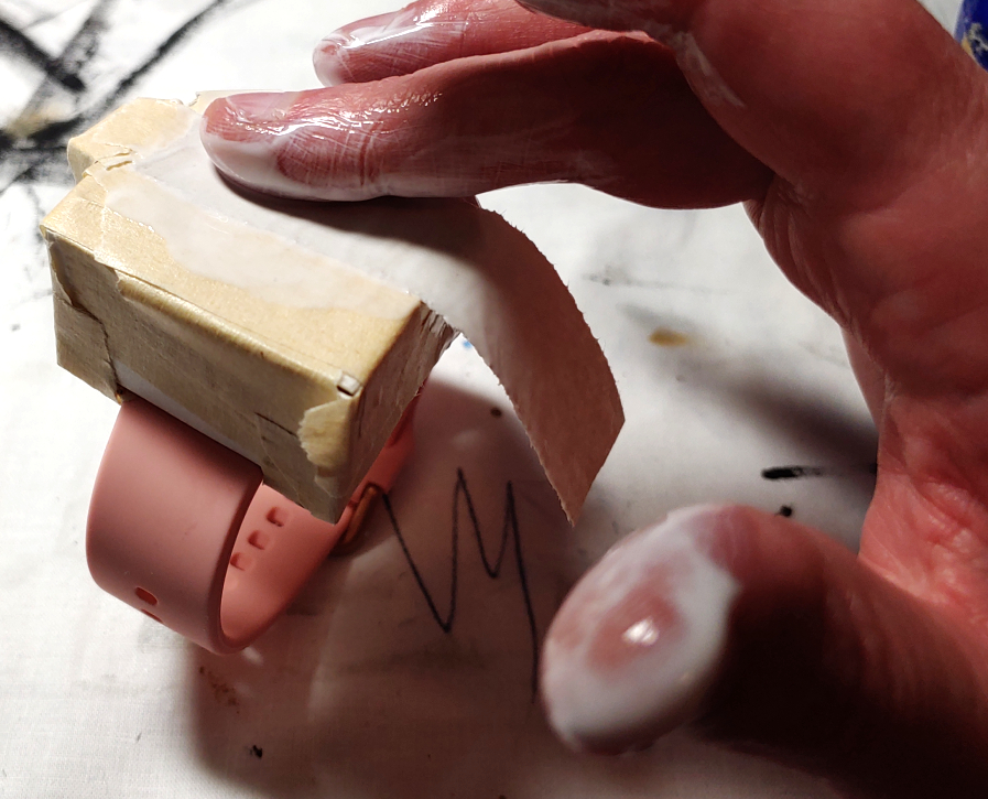 Rubbing a strip onto a small paper mache watch. Fingers coated in glue make sure this strip stays put and does not move