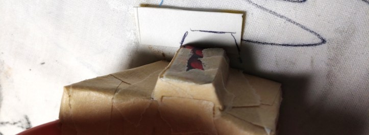 Tracing the side button cap onto a piece of food box. The Poketch is slightly displaced from the food box to show the pen marks.