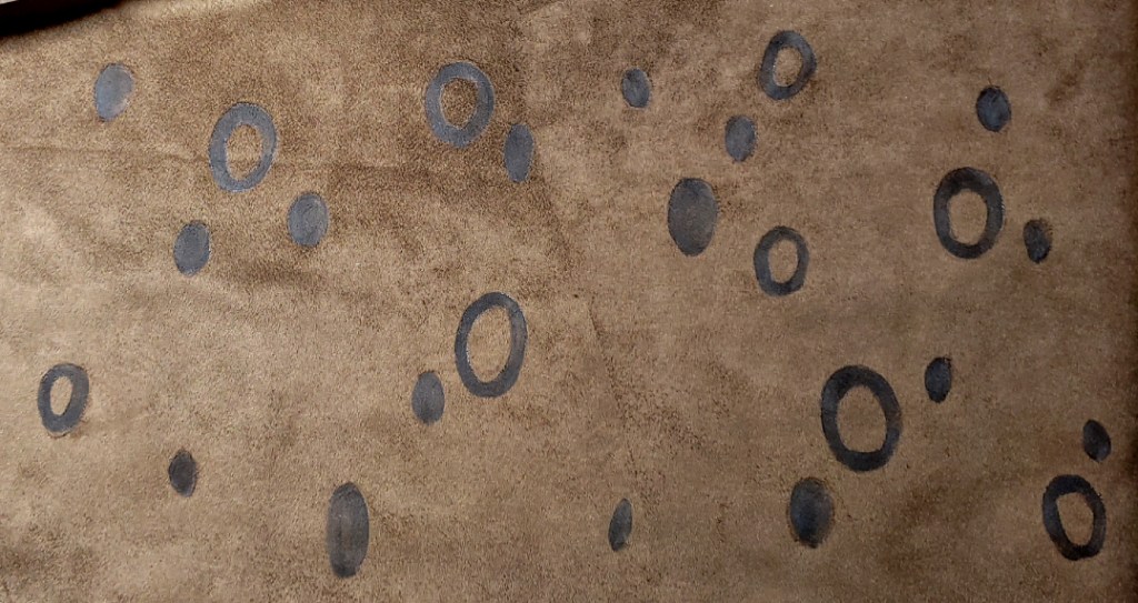 Cheetah-like spots painted on chocolate brown faux suede. The spots are irregular, some looking like a circle filled in and others looking like an O
