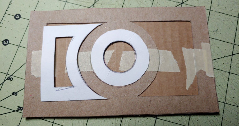 Using the discarded pattern pieces to determine placement of the raised details. White poster paper sits in the cutouts of the food box frame taped to the top.