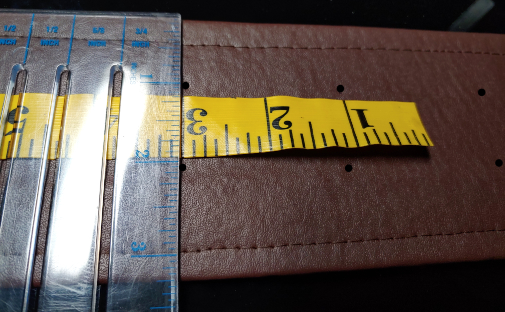 Using a tape measure and clear ruler to mark spacing. a yellow tape measure lays across the brown belt with a clear plastic ruler sitting perpendicular. black dots added to show pen markings