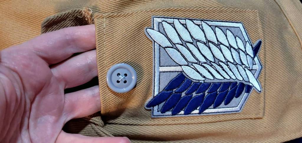 Gray button attached to the pocket over the Attack on Titan patch. My hand is in the pocket to show placement and pocket usability.