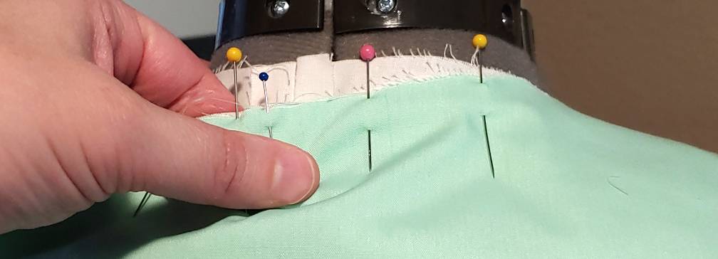 Pinning the collar to the shirt at center back. Several pins stick out of the light green collar as it's pinned to the white shirt underneath.