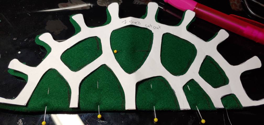 Green fabric fold pinned with yellow-head quilting pins while tracing and cutting out the details for a turtle shell using a white pattern piece. The pink in the back is my pen.