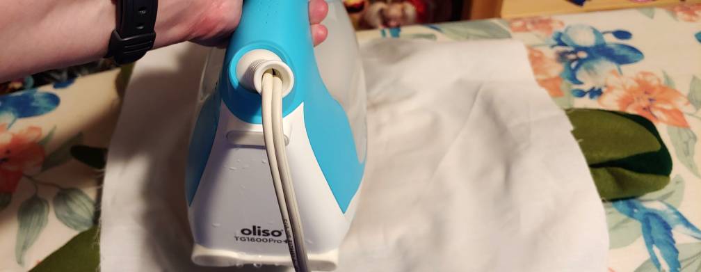 Pressing a turtle shell under a white muslin press cloth. The green turtle sticks out on either side of the white press cloth with a blue and white iron on top.