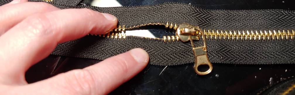 Showing the gap of teeth in the tape with the zipper pull slid nearly to the top. The gap is to the right of my middle finger with the light shining on my desk showing through it.