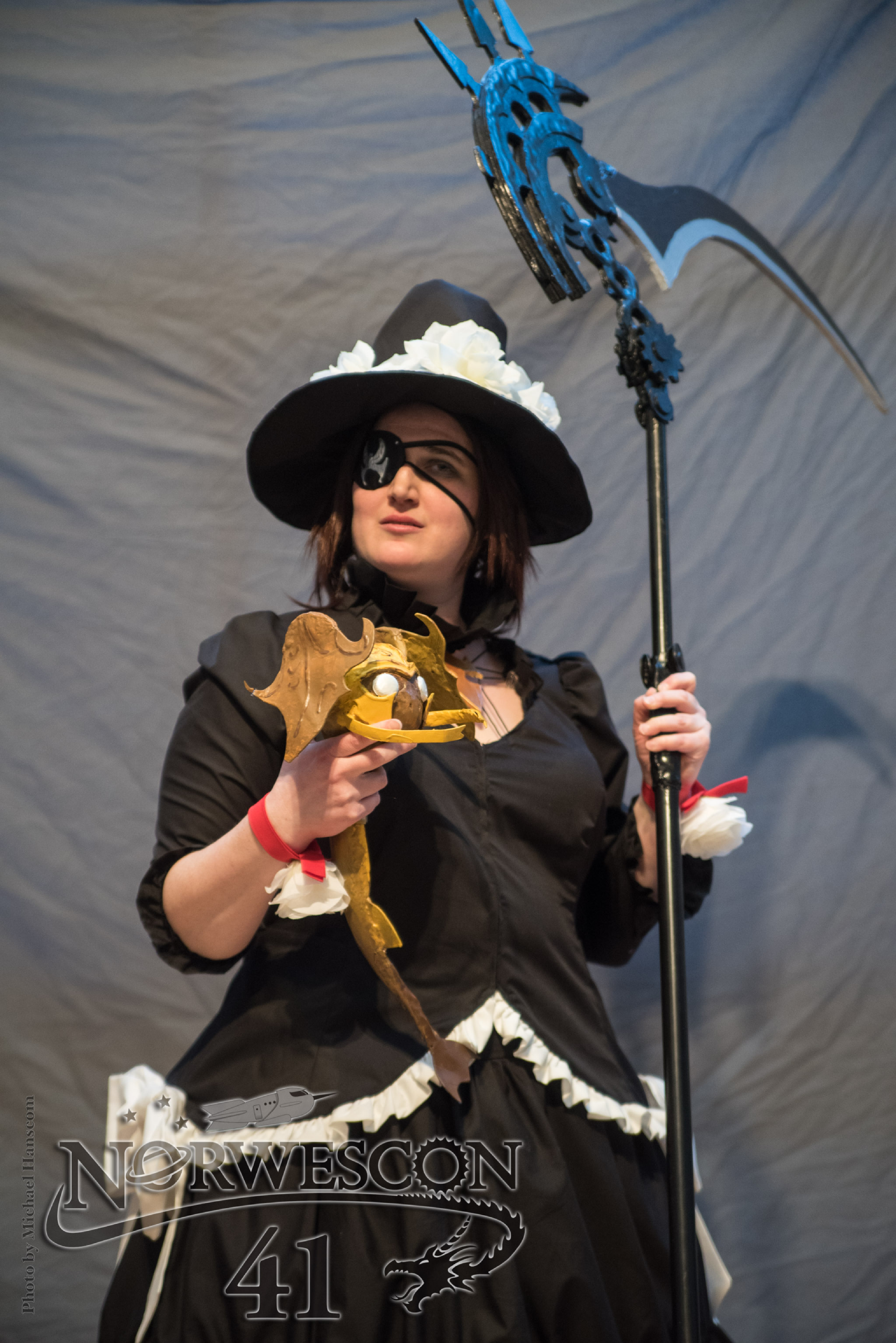 Edda holding Avere and her scythe in front of a gray curtain