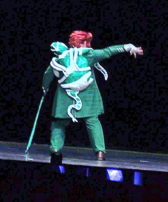 Back turned on a dark stage, hand outstretched to the right