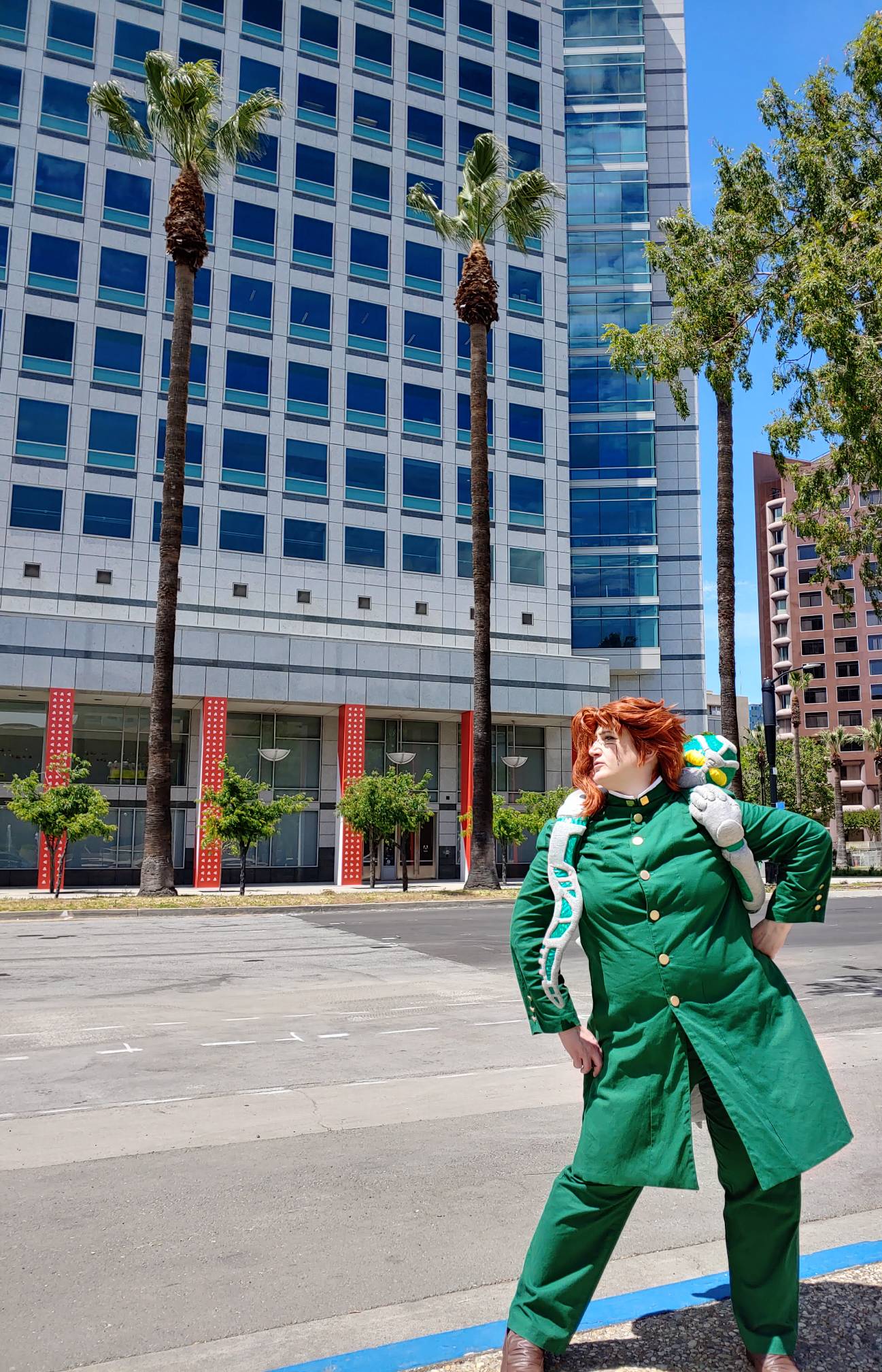 Kakyoin with hand on hip with Hierophant Green wrapped around the shoulders, posing in front of palm trees and a skyscraper.