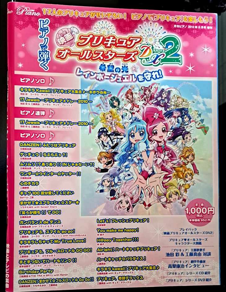 Front cover featuring fireworks and pink sparkles on the top border with "PreCure All Stars DX2" Written in Japanese. The left and bottom text are all the songs included inside (refer to song list above for translations). On the right is the DX2 artwork with Cure Blossom and Cure  Marine in the center with all the other Cures around in a circle.