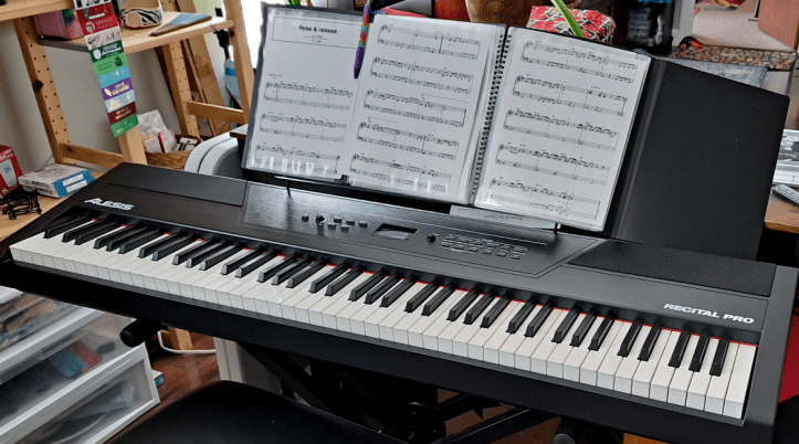 Electronic piano set up against a desk with 3 sheets of music sitting on the stand.