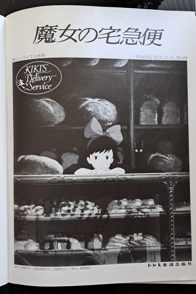 Title page with "Kiki's Delivery Service written in Japanese across the top. The image is Kiki leaning against the bread shop counter, depressed, with Jiji sitting next to her. A stamp of "Kiki's Delivery Service" is in the upper left.