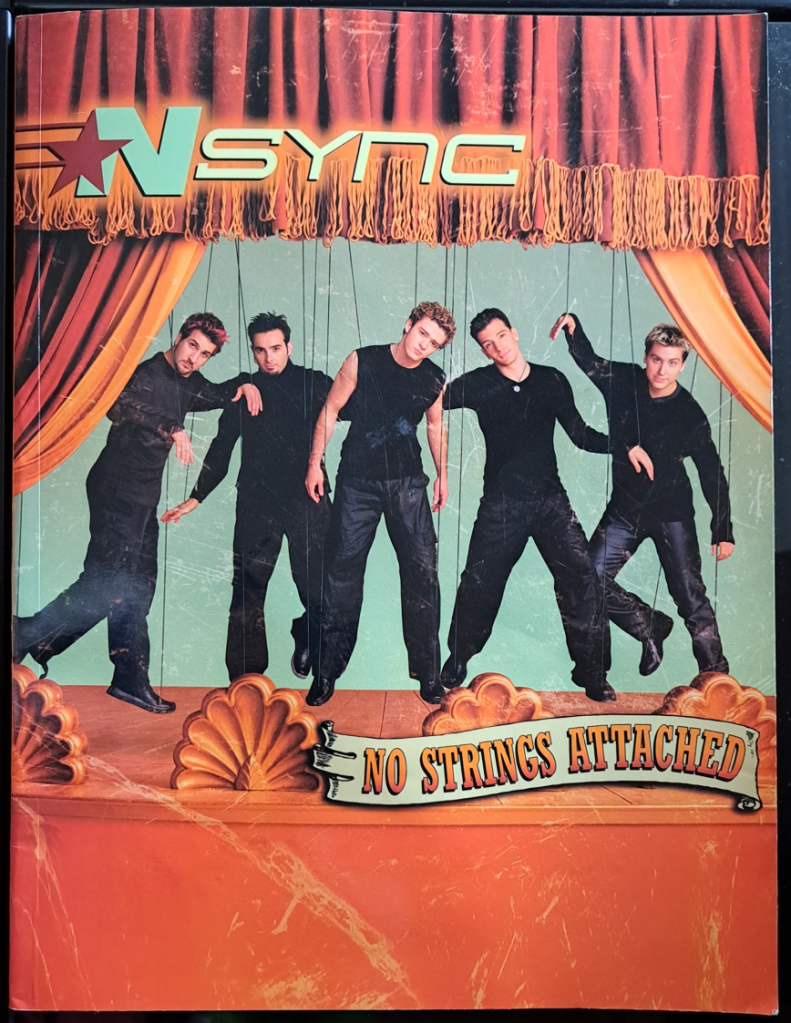Orange-red front cover with curtains pulled to the side. NSYNC is in the middle in black with strings to look like marionettes. From left to right: Joey, Chris, Justin, JC, and Lance