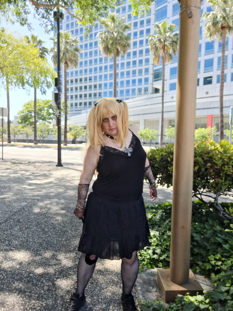 Misa giving a death stare, standing a bit crooked, in a park with palm trees and a tall building in the background