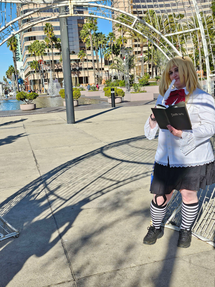 Misa standing, holding the death note and deviously holding a feather pen to her lips